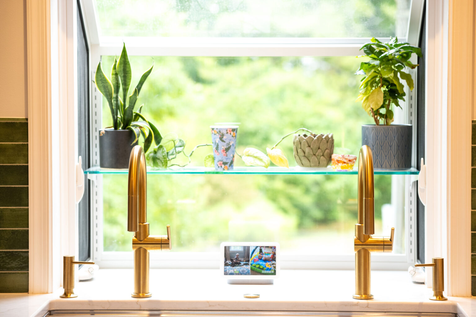 Image by Lesnick Photo Two gold kitchen faucets under a bright window with plants, a floral cup, and a digital display on a glass shelf. Greenery is visible outside.