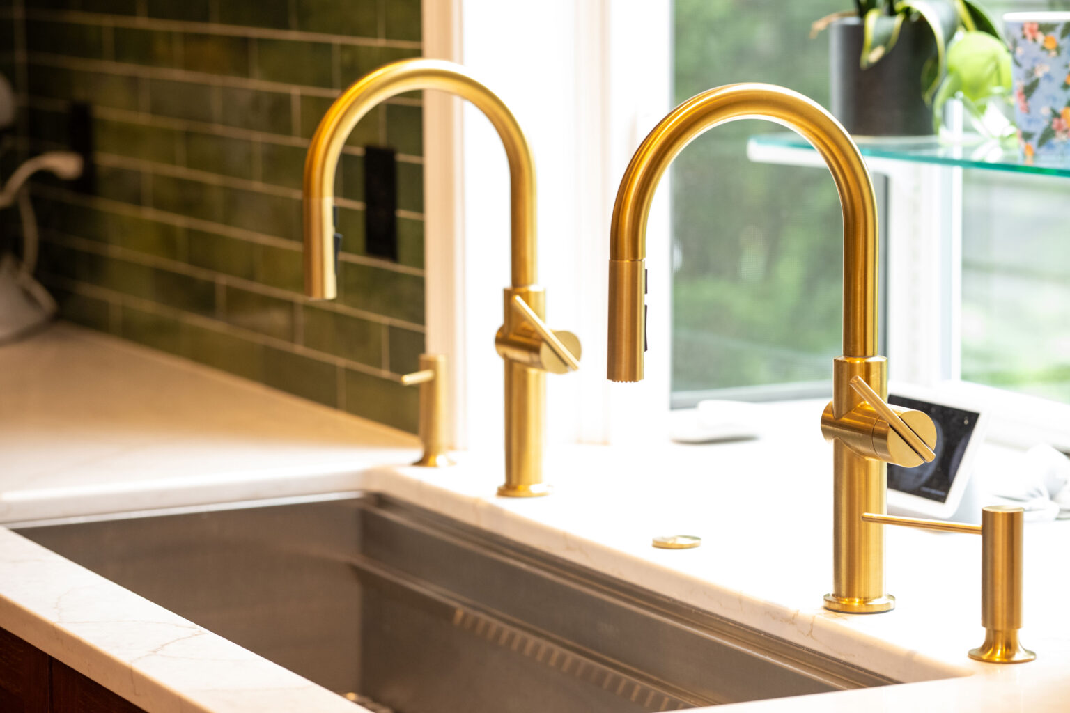 Image by Lesnick Photo A modern kitchen sink with two gold faucets against a green tiled wall, set beneath a window.