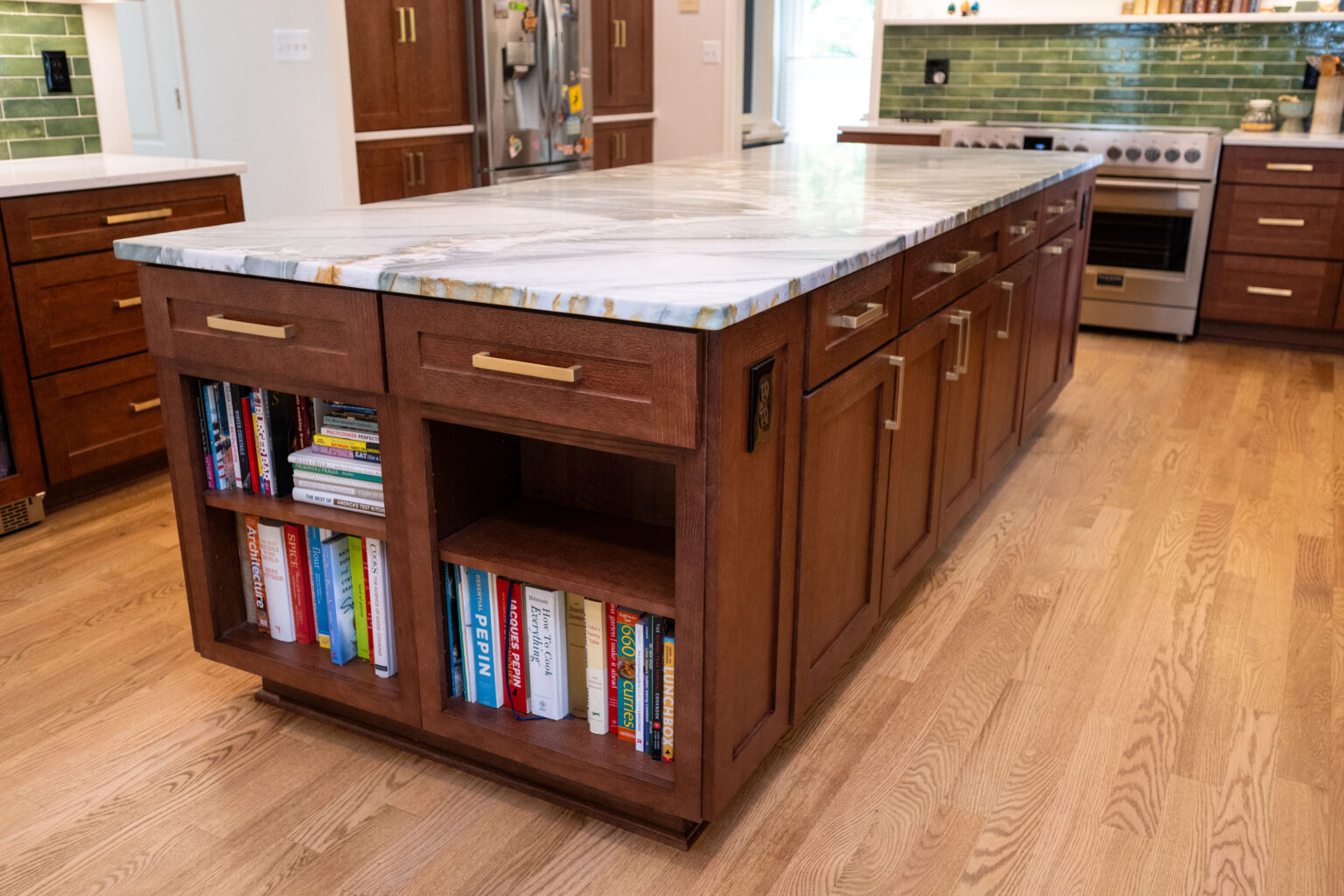 Image by Lesnick Photo Wooden kitchen island with marble countertop and bookshelves containing various books. Green tiled backsplash is visible in the background.