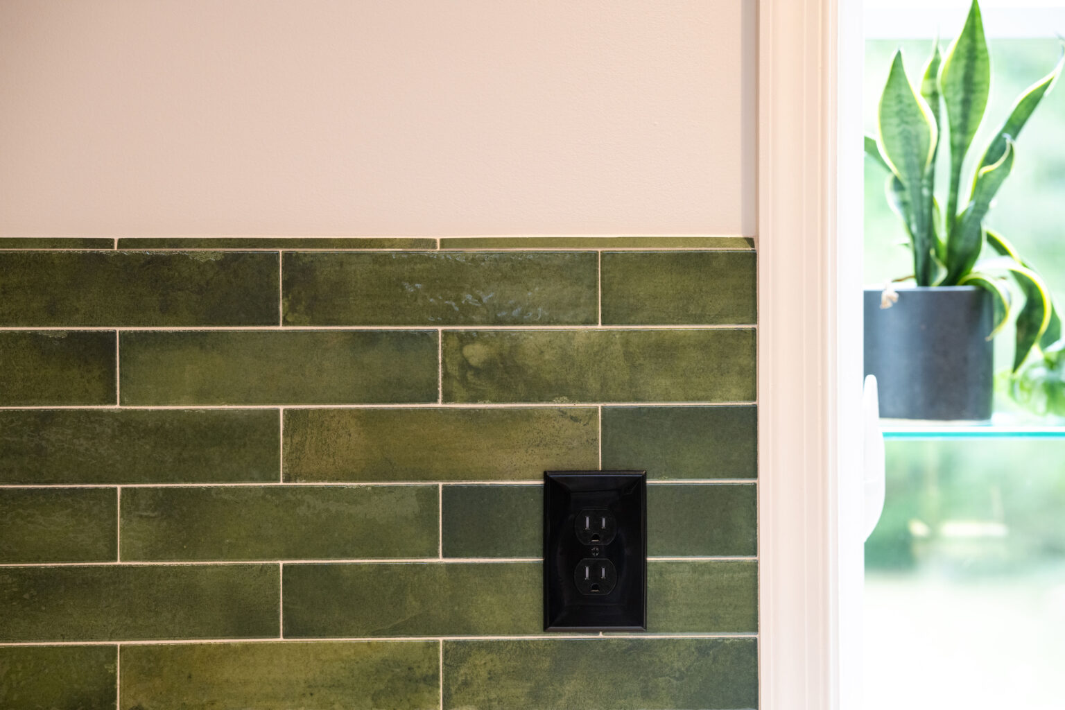 Image by Lesnick Photo Green tiled wall with a dark electrical outlet, adjacent to a window with a potted snake plant on the sill.