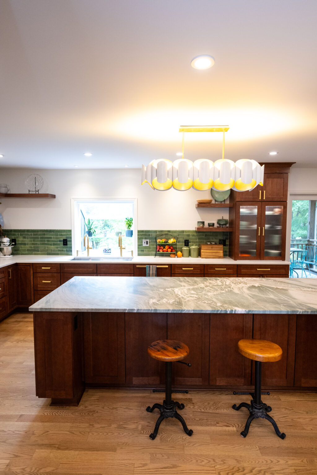 Image by Lesnick Photo Modern kitchen with wooden cabinets, a marble island, two round stools, and a unique light fixture. Green subway tiles line the backsplash. A window with a view is in the background.