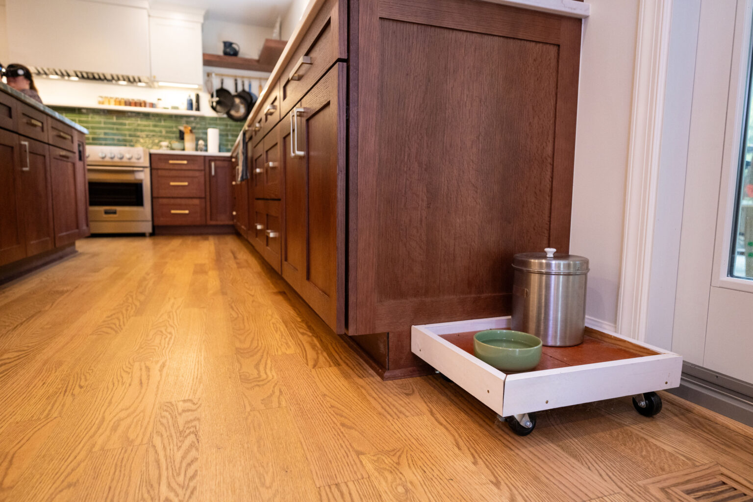Image by Lesnick Photo A kitchen with wooden cabinets and a hardwood floor, featuring a pull-out tray with a green bowl and a metal container.