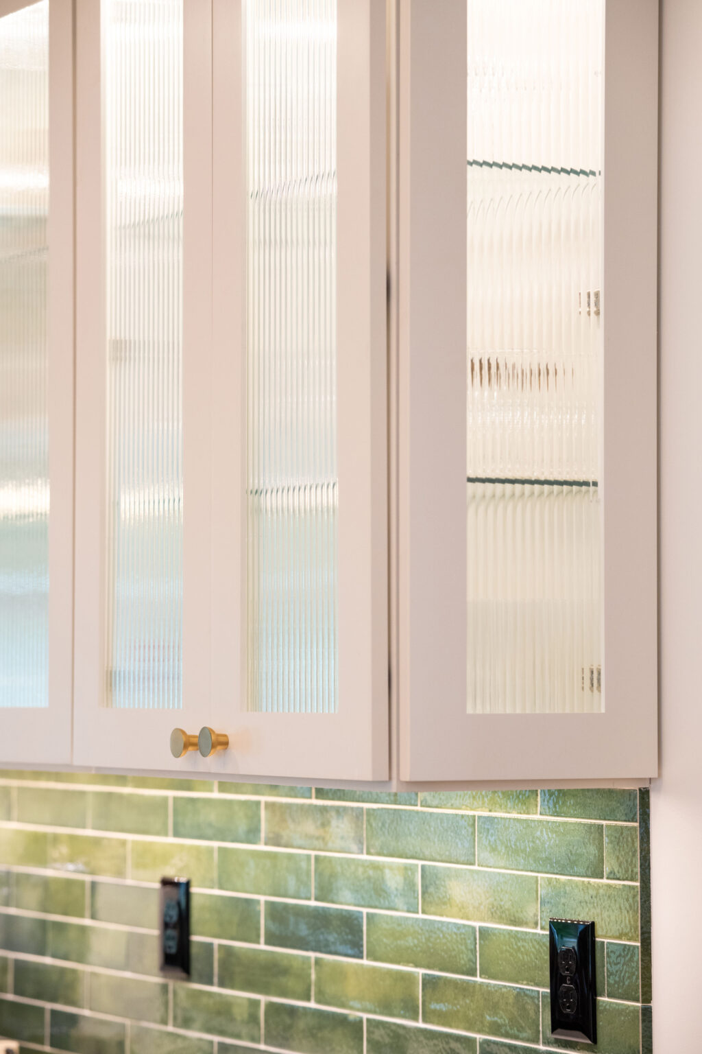 Image by Lesnick Photo Kitchen cabinets with fluted glass doors above a backsplash of green subway tiles and black wall outlets.