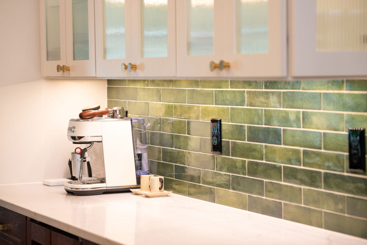 Image by Lesnick Photo A modern kitchen counter with a coffee machine and a cup, set against a green tiled backsplash and white cabinets with gold handles.