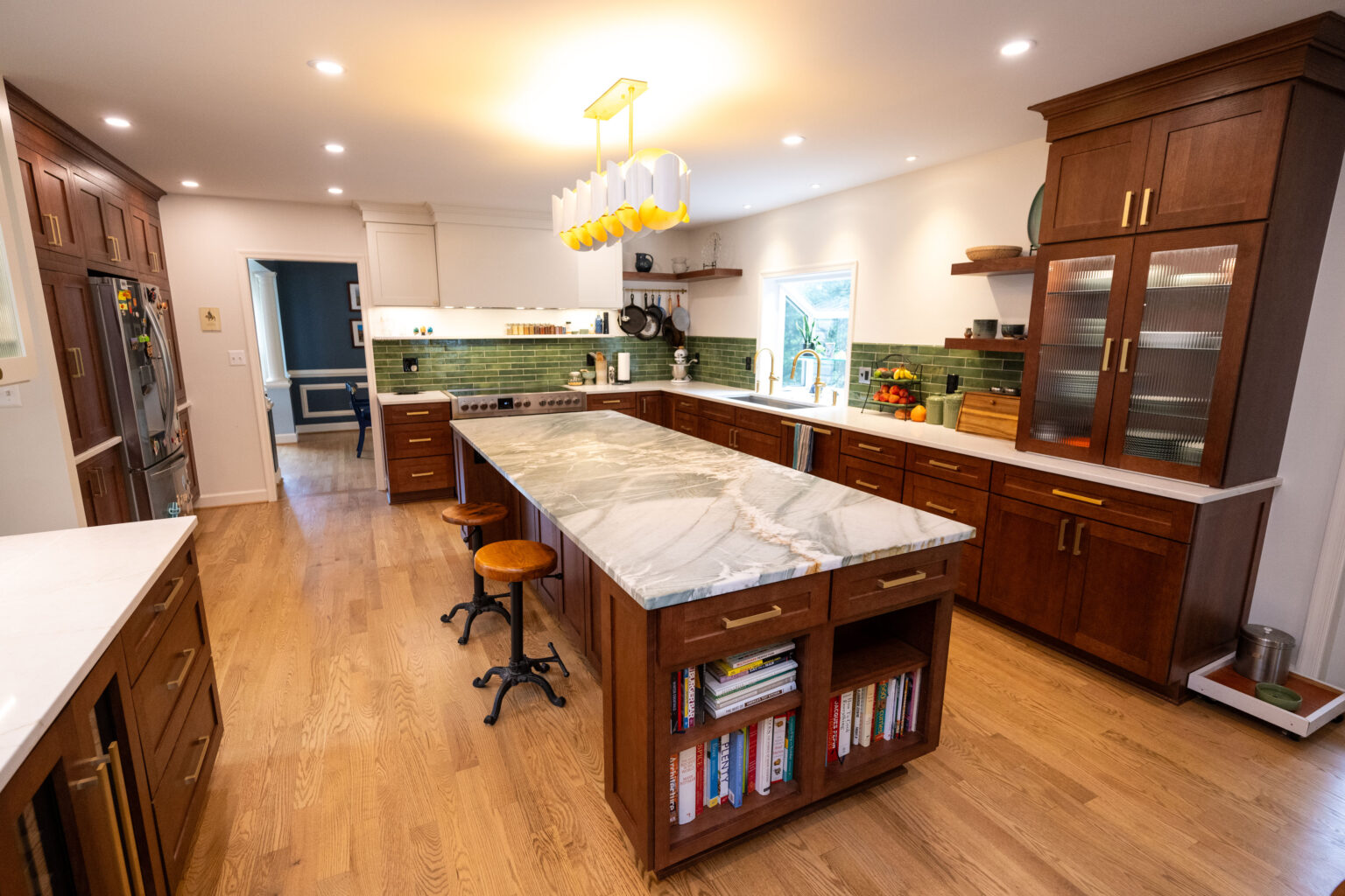 Image by Lesnick Photo Modern kitchen with wooden cabinets and a large marble island. Green tile backsplash, pendant lighting, stools, hardwood floor, and books stored on the island's shelves.