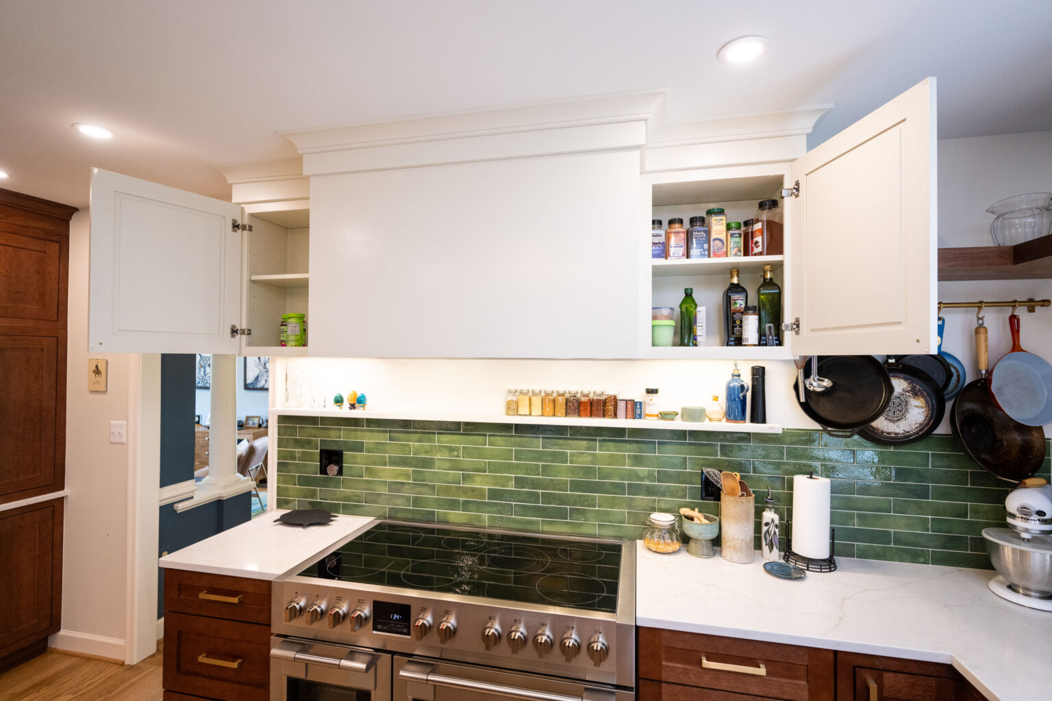 Image by Lesnick Photo Modern kitchen with open cabinets revealing spices and oils. Green tile backsplash, stainless steel stove, and cookware hanging on the wall.