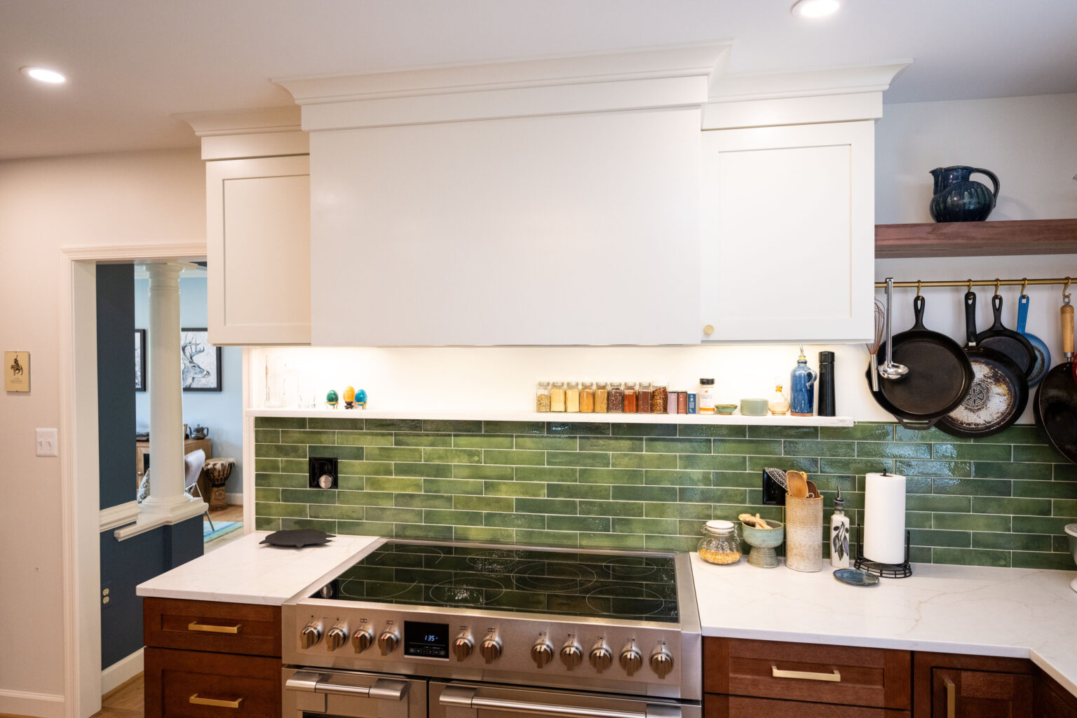 Image by Lesnick Photo Modern kitchen with a white range hood, green tile backsplash, and stainless steel stove. Wooden cabinets below and various kitchen items on the counter and shelves above.