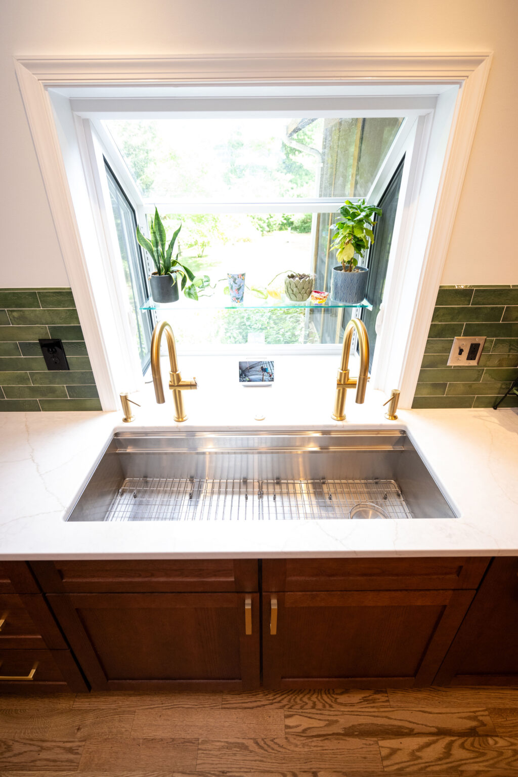 Image by Lesnick Photo A stainless steel kitchen sink with brass faucets is set beneath an open window. Green tiled backsplash. Potted plants are placed on the windowsill.