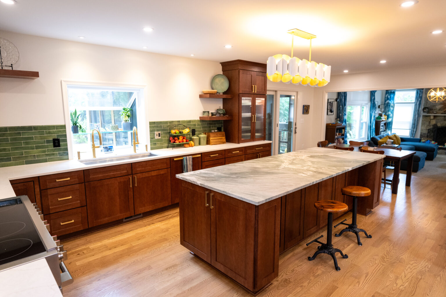 Image by Lesnick Photo Spacious kitchen with wooden cabinets, a large marble island, and modern pendant lighting. Green tile backsplash and hardwood flooring add warmth. Sunlight streams through a window and glass door.