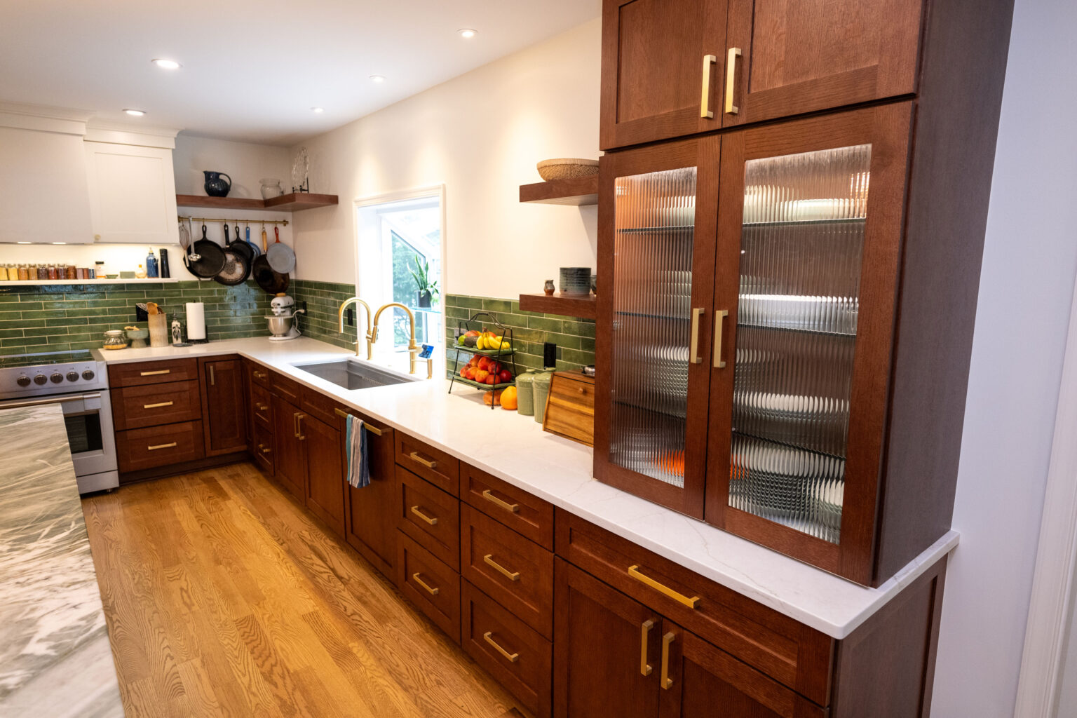 Image by Lesnick Photo A modern kitchen with wooden cabinets, a white countertop, and green tile backsplash. A stove is on the left, and a sink is centered under a window. Shelves hold dishes and cookware.