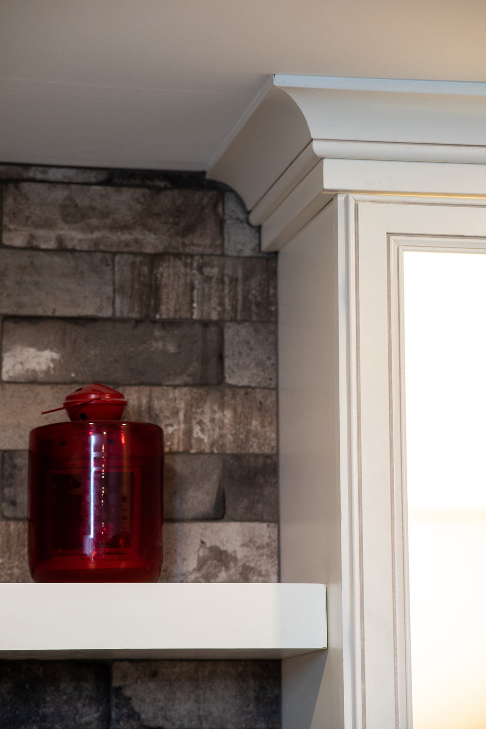 A red glass jar sits on a white shelf against a gray brick wall under a cabinet with crown molding.