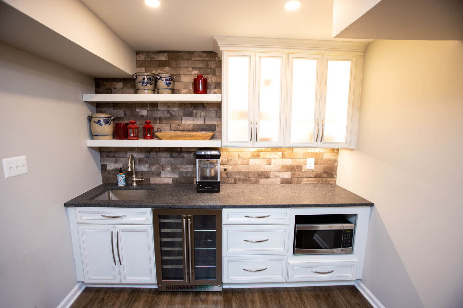 A small kitchen area with white cabinets, a stone backsplash, open shelves with jars, a sink, a coffee maker, wine cooler, and a microwave. The countertop is dark and floor is wooden.
