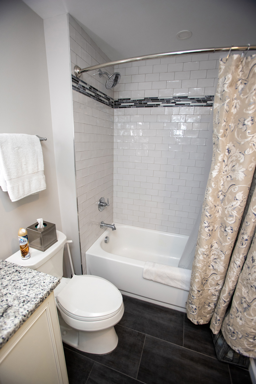Modern bathroom with a white bathtub, tiled walls, a patterned shower curtain, a toilet, and a granite countertop with tissue box and air freshener.