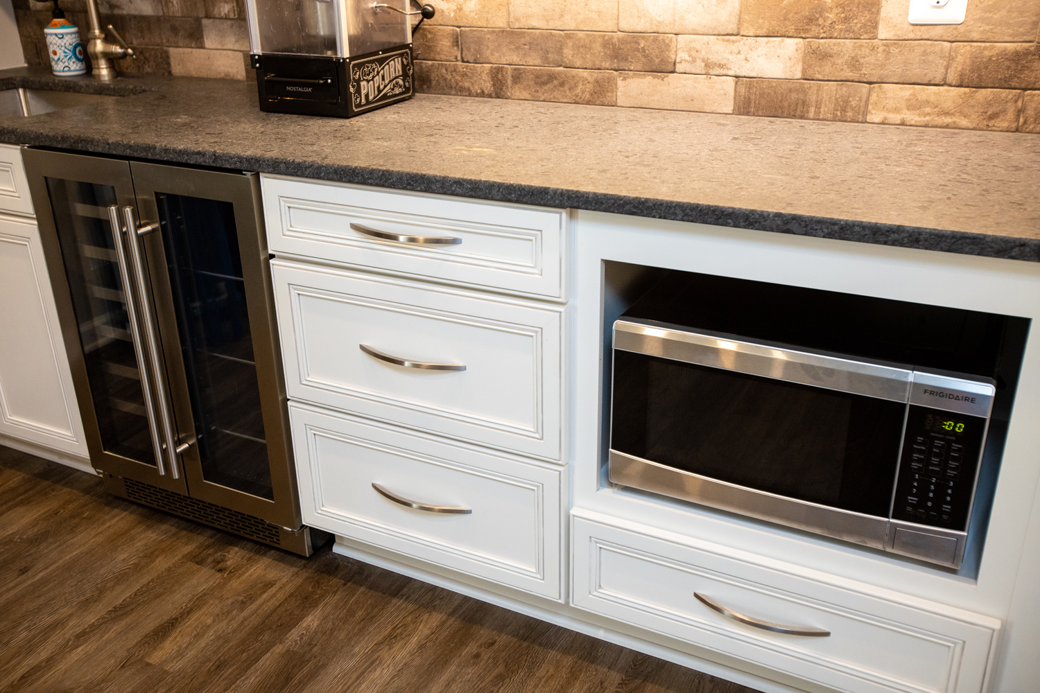Kitchen countertop with white cabinets, a stainless steel microwave, and a wine cooler. Wood flooring and a stone backsplash complete the setup.