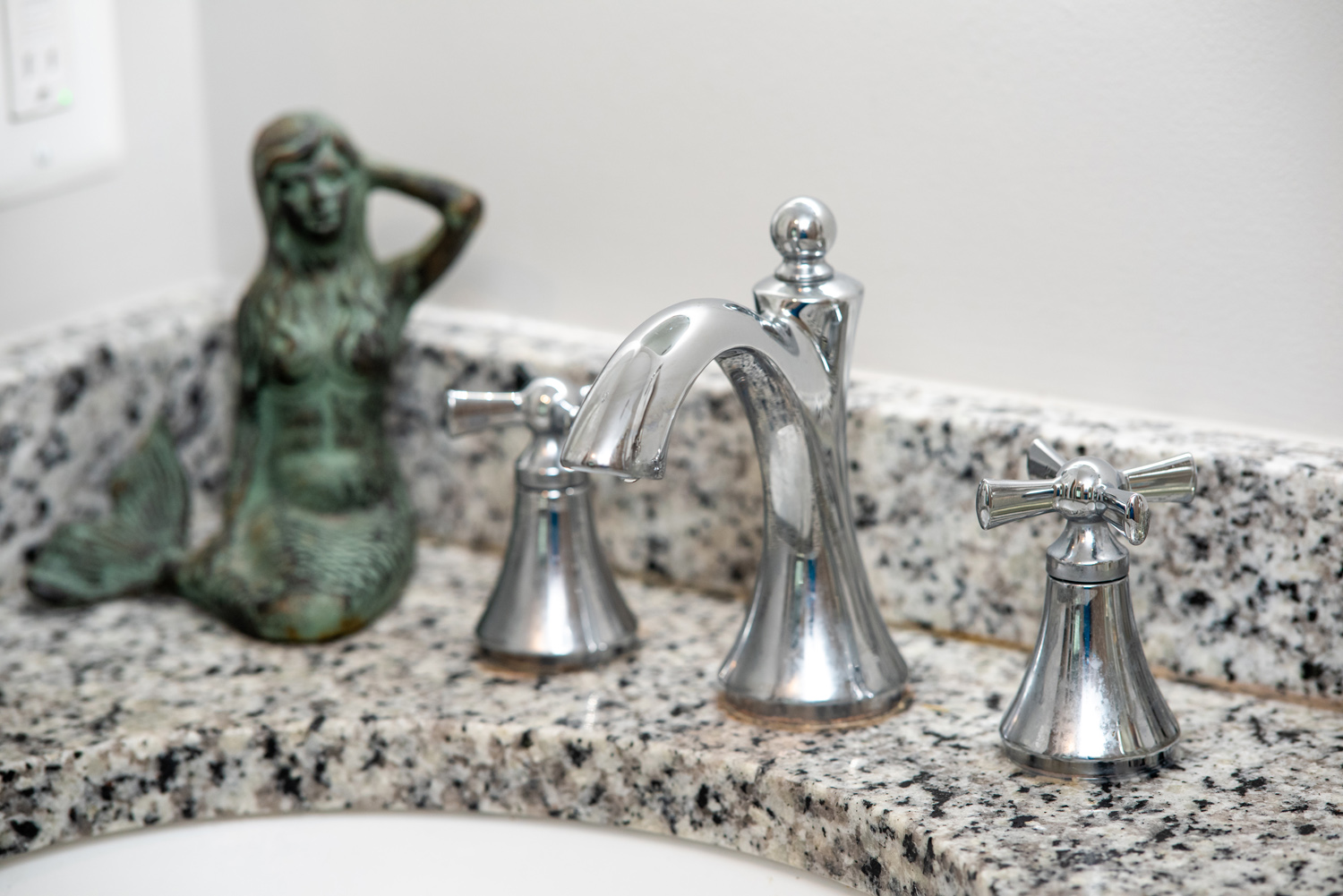 A bathroom sink faucet with two handles on a granite countertop, accompanied by a bronze mermaid sculpture.