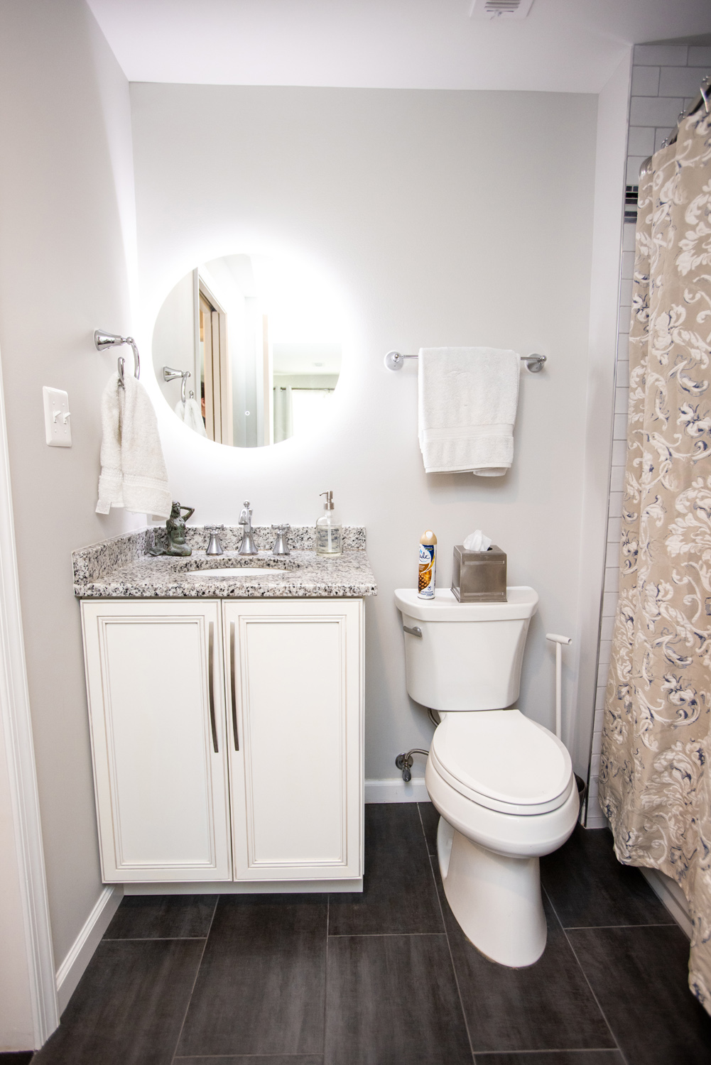 A small bathroom with a granite countertop, white vanity, circular mirror, toilet, towel rack, and a shower with a patterned curtain.