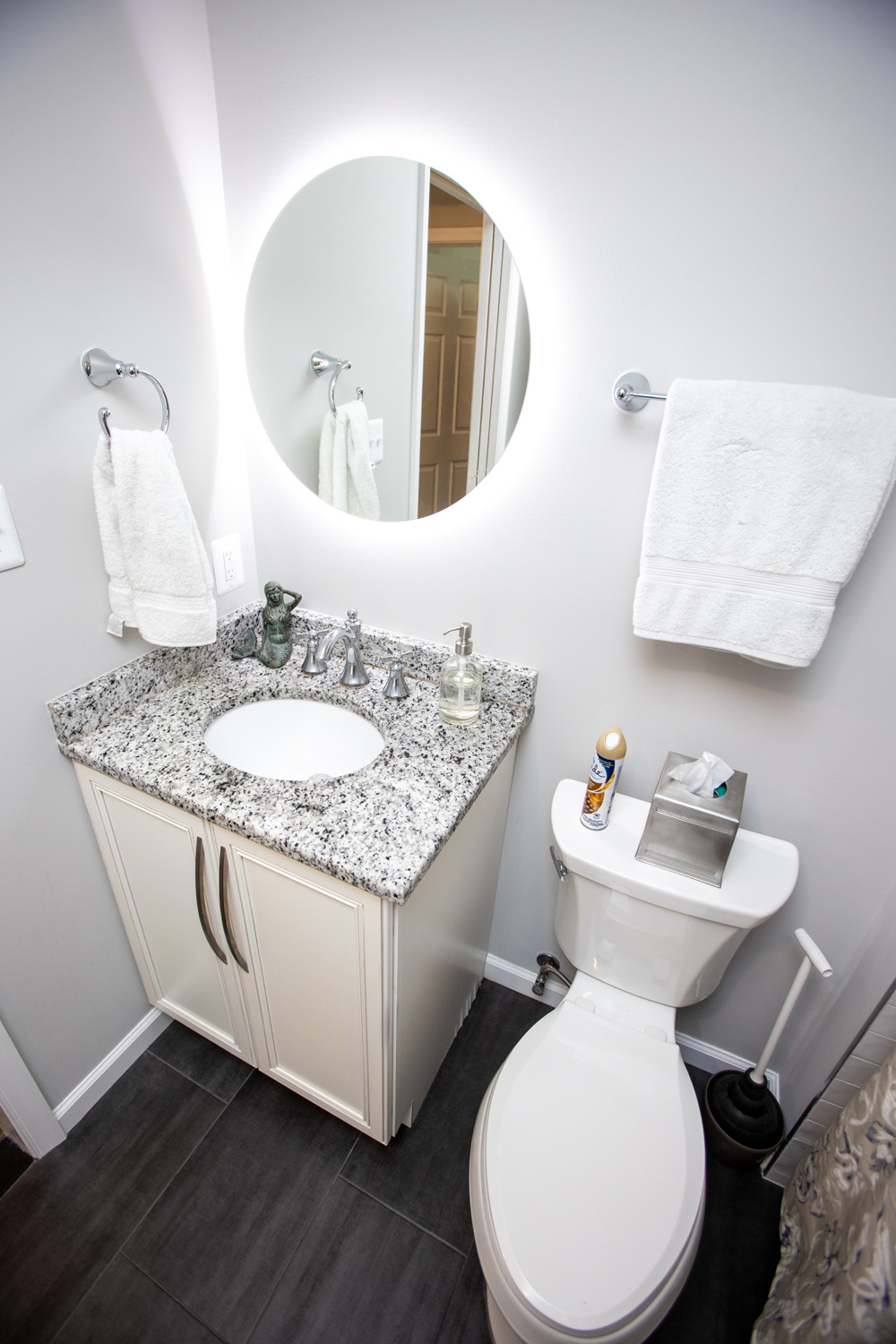 Bathroom with a granite countertop sink, a round backlit mirror, and a toilet. White towels hang on the wall, and a tissue box and air freshener are on the toilet tank.