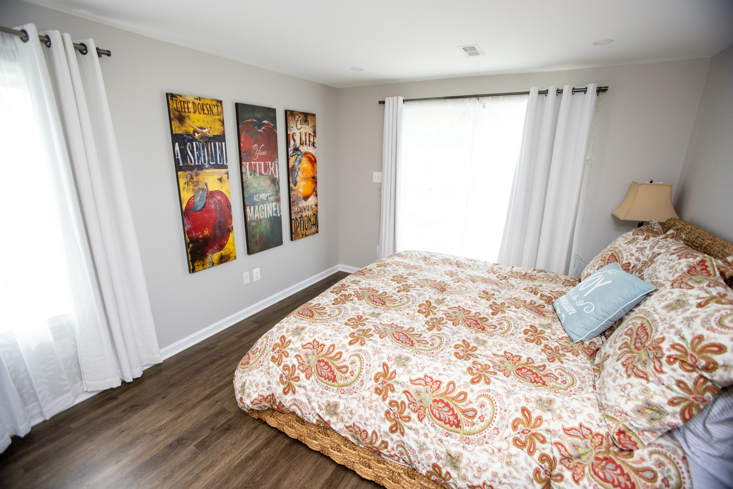 Bedroom with a patterned bedspread, artwork on the wall, sheer privacy curtains, and a wooden floor.