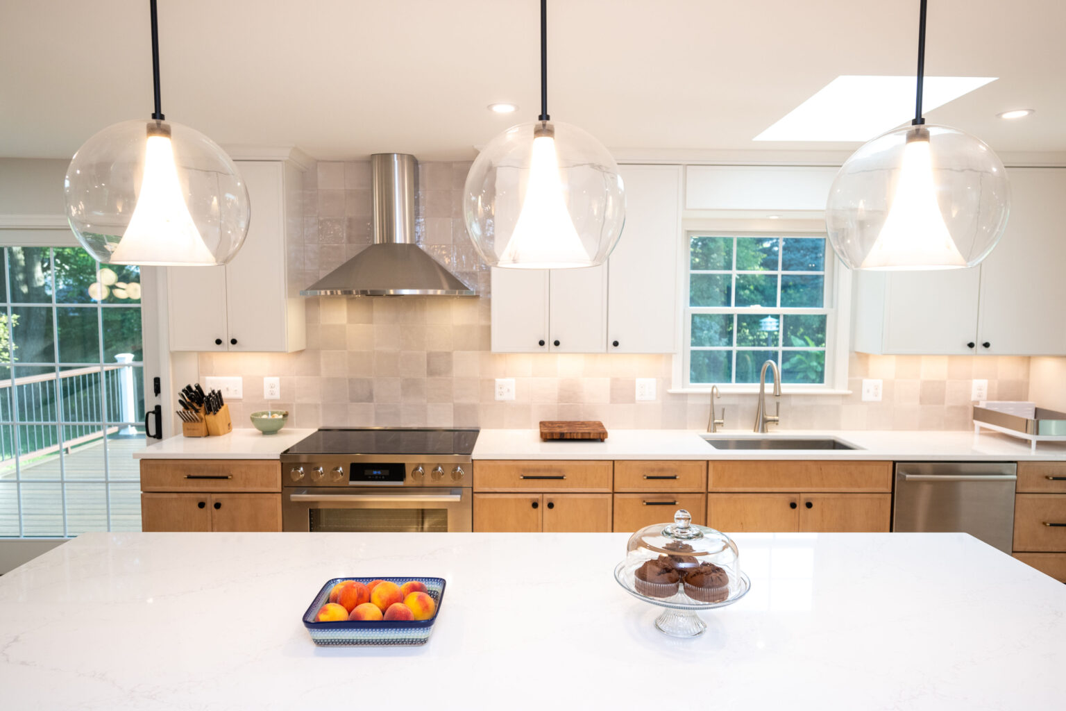 Modern kitchen with a marble island, pendant lights, wooden cabinets, and stainless steel appliances. A fruit bowl and cake stand are on the counter, with a view of a patio through sliding doors.