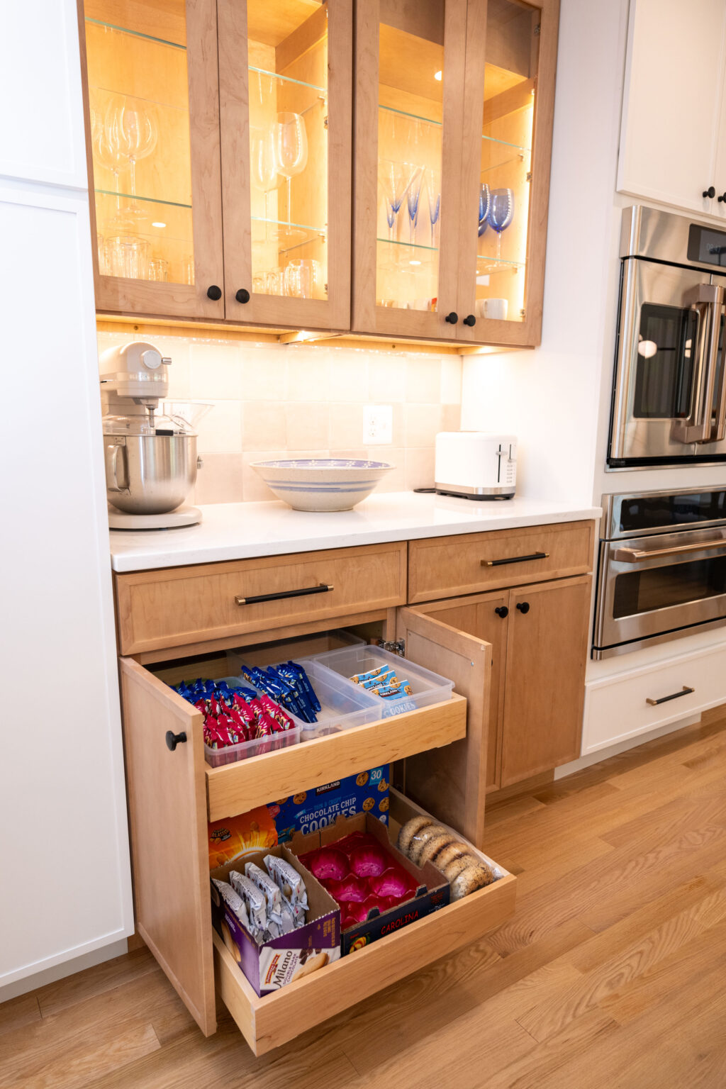Kitchen with wooden cabinets, an open drawer displaying snack packets, a stand mixer, a bowl, a toaster, and glassware in glass-front cabinets above.
