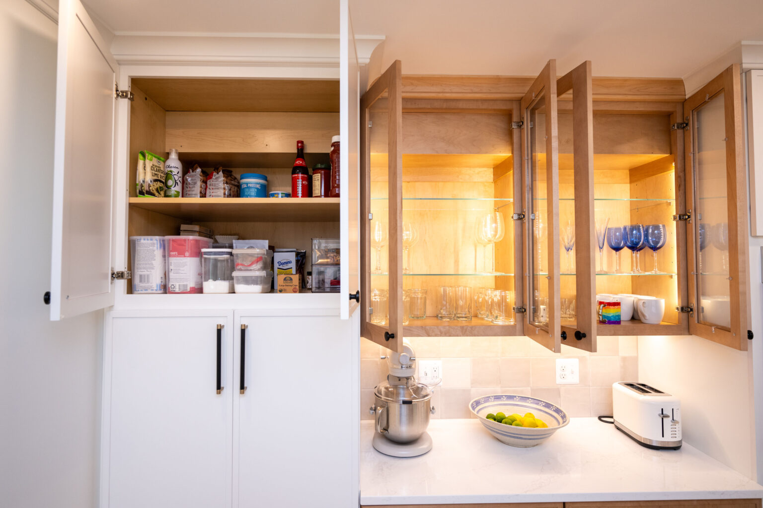 Kitchen shelves with open cabinets; left contains groceries and containers, right displays glasses. Countertop holds a mixer, fruit bowl, and toaster.