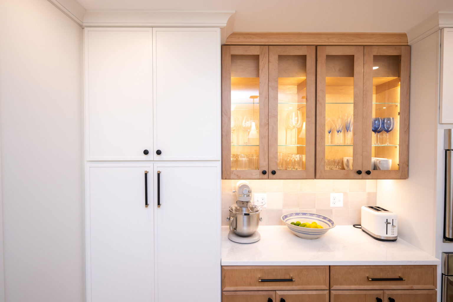 A kitchen with white cabinets, wooden upper cabinets with glass doors, a countertop with a stand mixer, a fruit bowl, and a toaster.