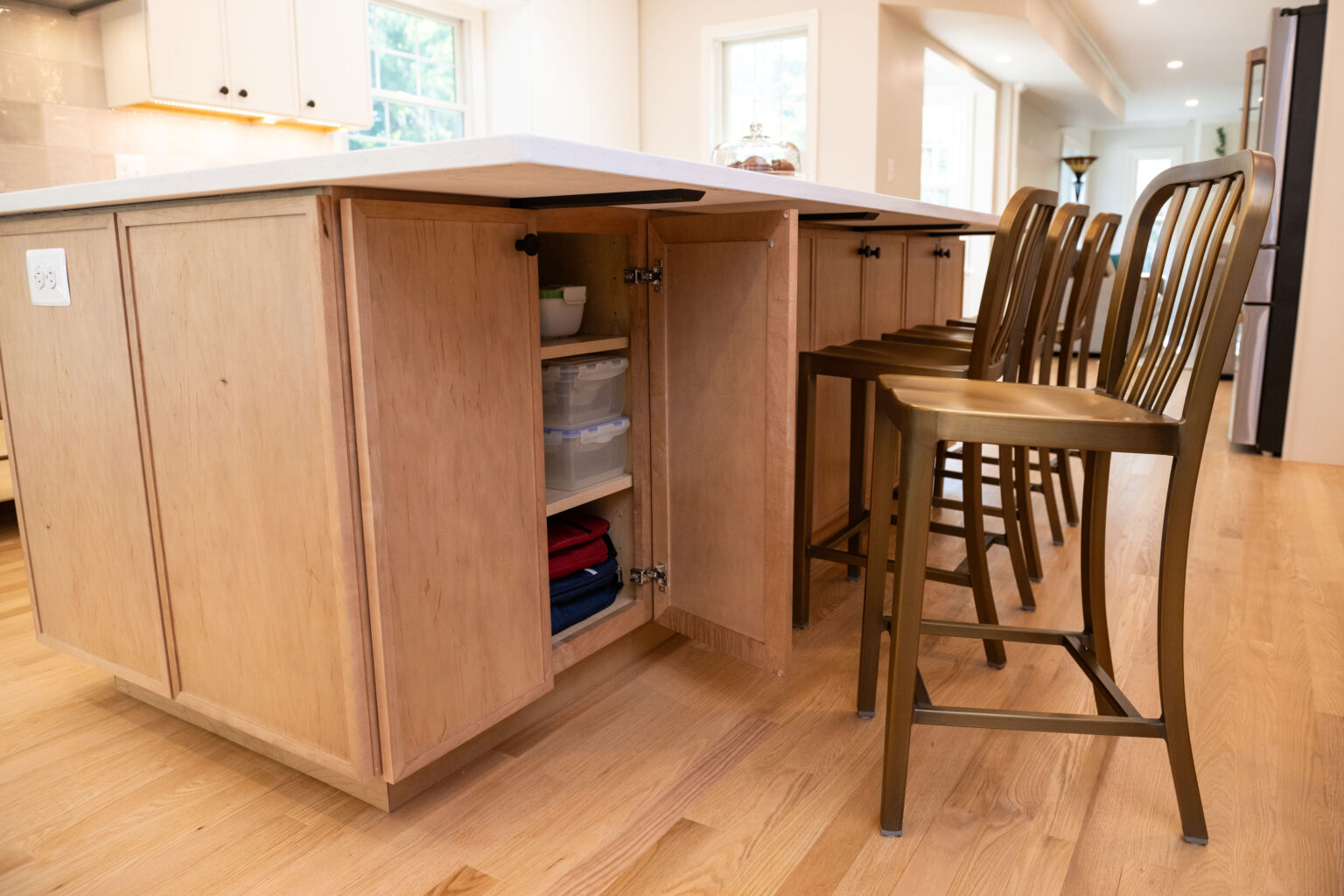 Kitchen island with light wood cabinets and open storage shelves. Four metal barstools are lined up on the right side. Bright and spacious room with large windows.