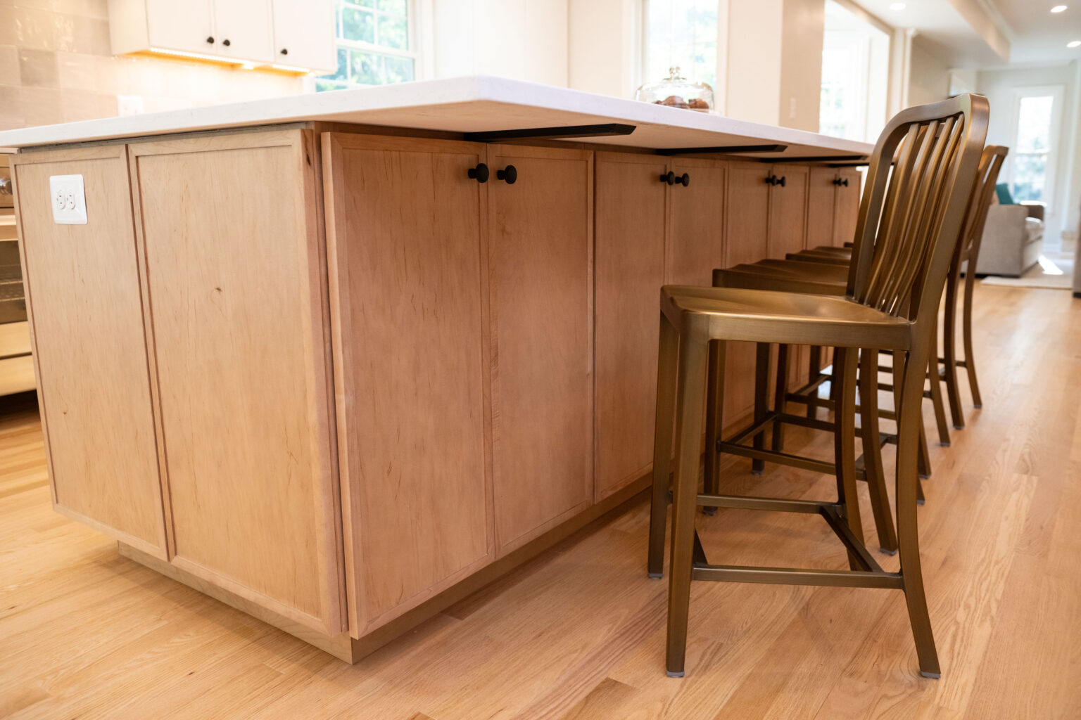 Wooden kitchen island with white countertop and four bronze stools on a light hardwood floor, illuminated by natural light from nearby windows.