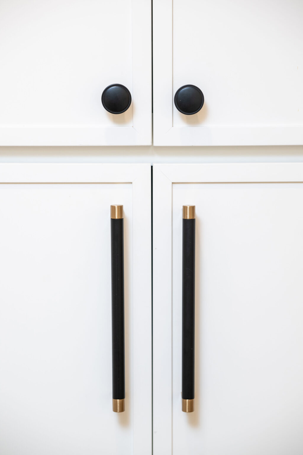 White cabinet doors with black circular knobs on the top pair and long black handles with gold accents on the bottom pair.
