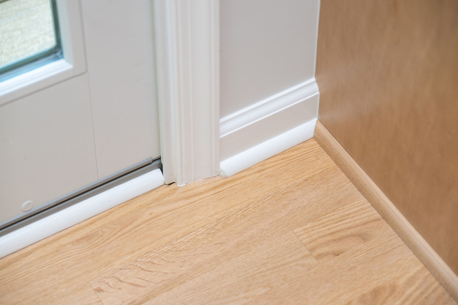 Close-up of the bottom corner of a door frame with white trim, adjacent to a light wood floor and a wooden wall panel.