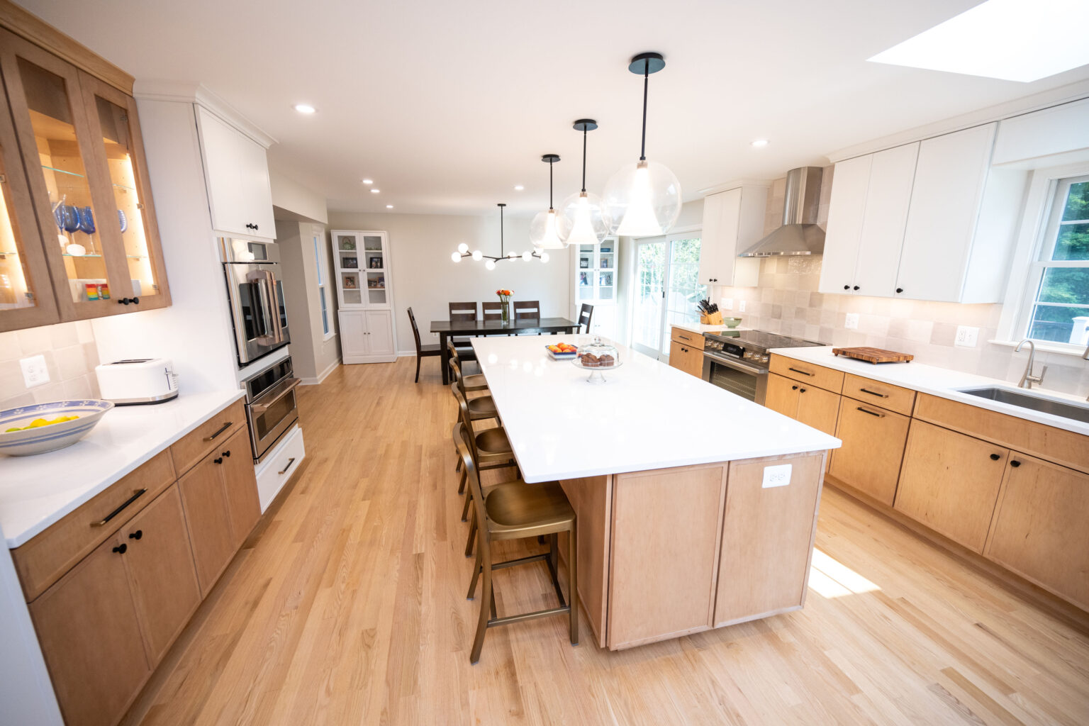 Spacious kitchen with wooden cabinets, a large island with stools, pendant lights, and stainless steel appliances. Dining area visible in the background.