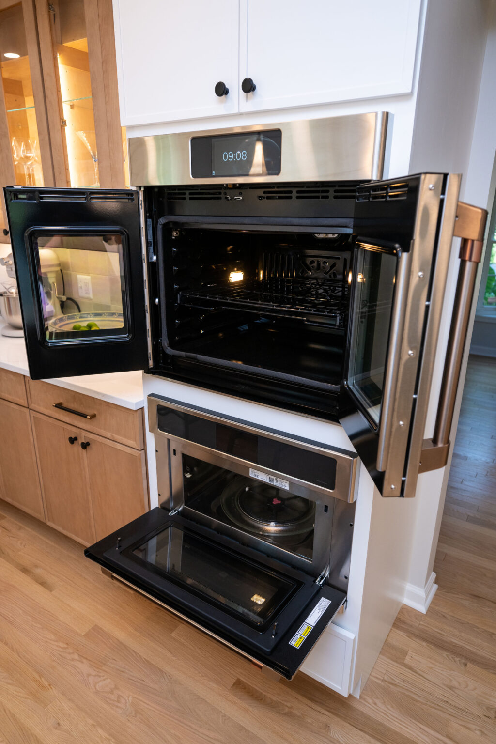 A modern kitchen with an open microwave and an open wall oven built into white cabinets on a wood floor.