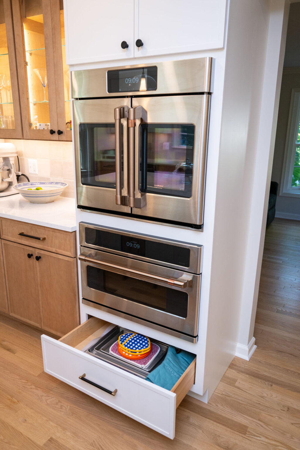 A modern kitchen features a stainless steel double oven with digital displays, wooden cabinets, a white drawer partially open showing a colorful dish, and hardwood flooring.