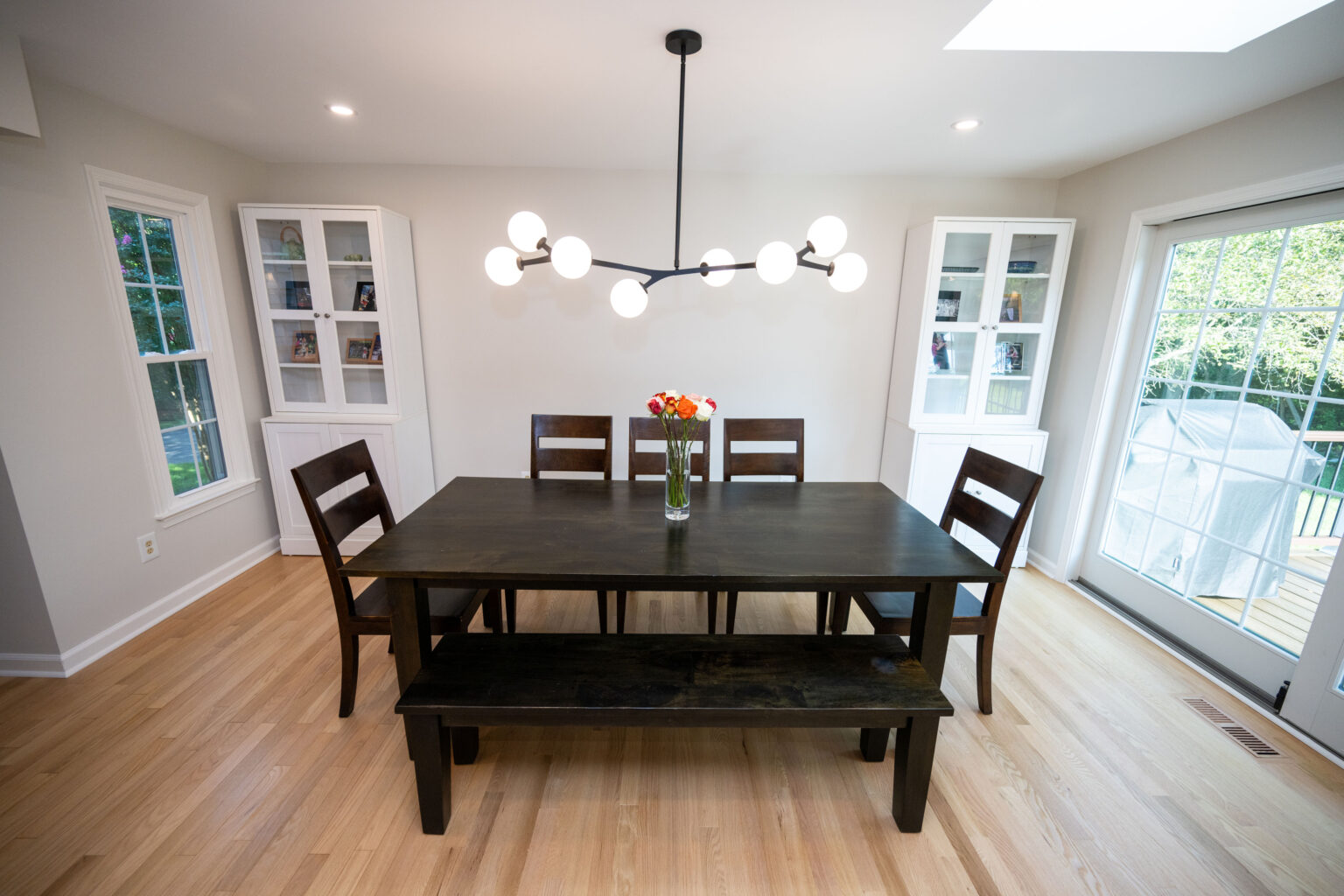 Dining room with a dark wood table, chairs, and bench. A modern chandelier hangs above, and there are glass double doors and built-in shelves on either side. A vase with flowers is on the table.