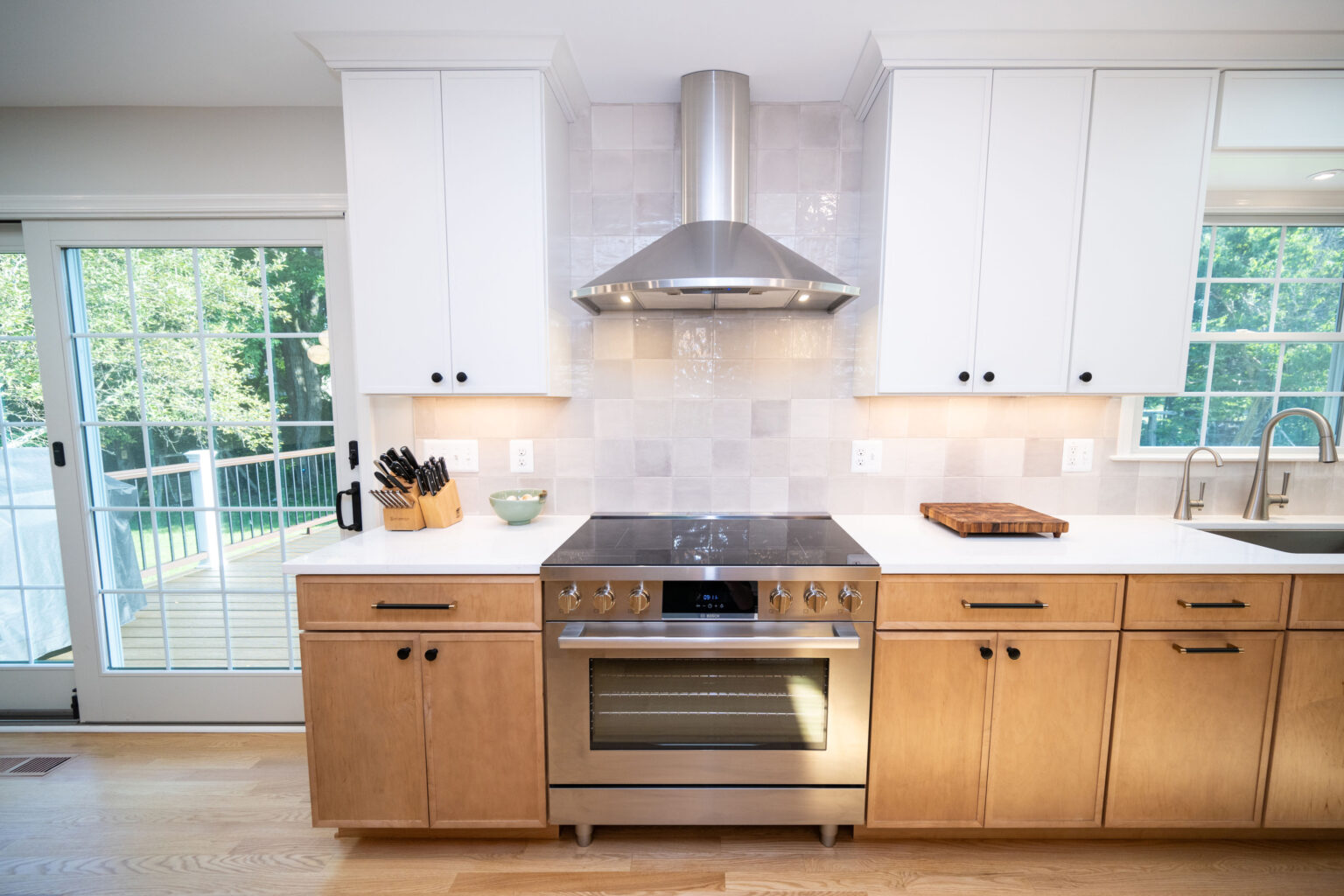 Modern kitchen with a stainless steel oven and range hood, white upper cabinets, wooden lower cabinets, and a view of trees through a sliding glass door.