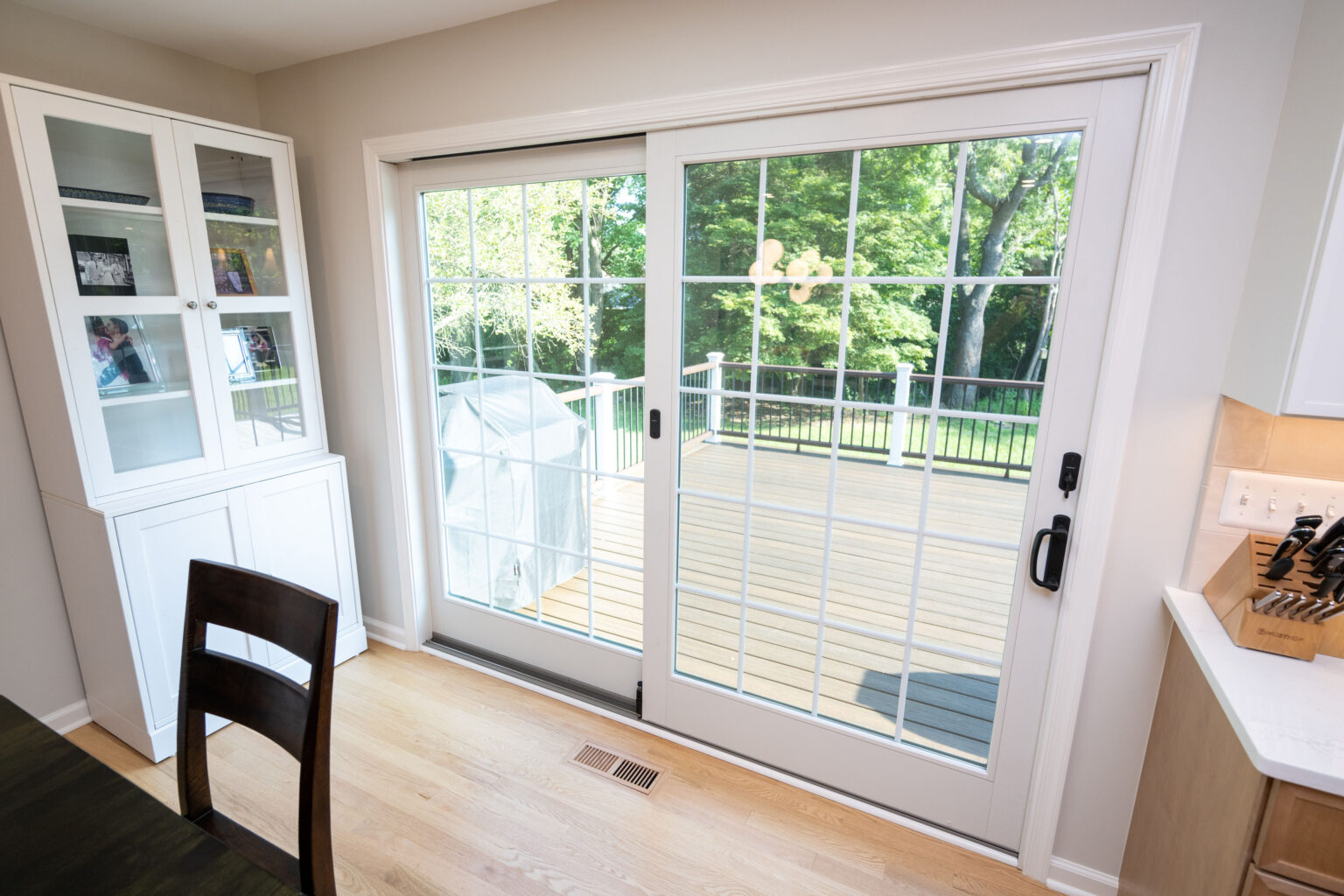 Glass sliding doors open to a wooden deck with a view of green trees. Indoors, there's a chair, a countertop with a knife block, and a white cabinet.