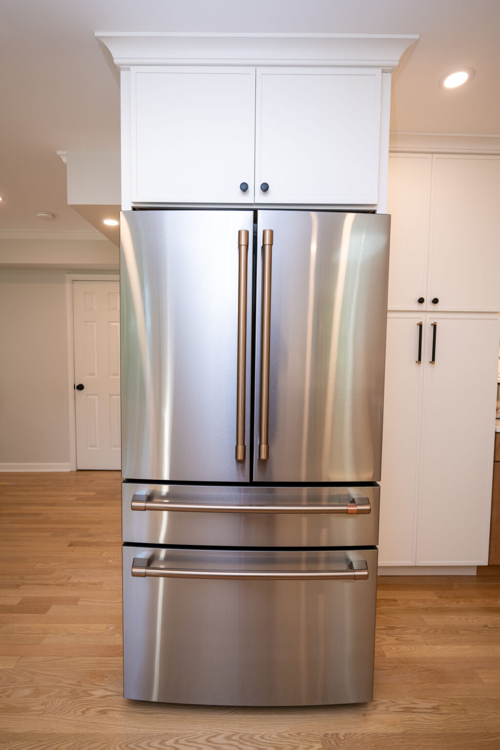 A stainless steel, French door refrigerator with two bottom freezer drawers, positioned in a modern kitchen with light wood flooring.