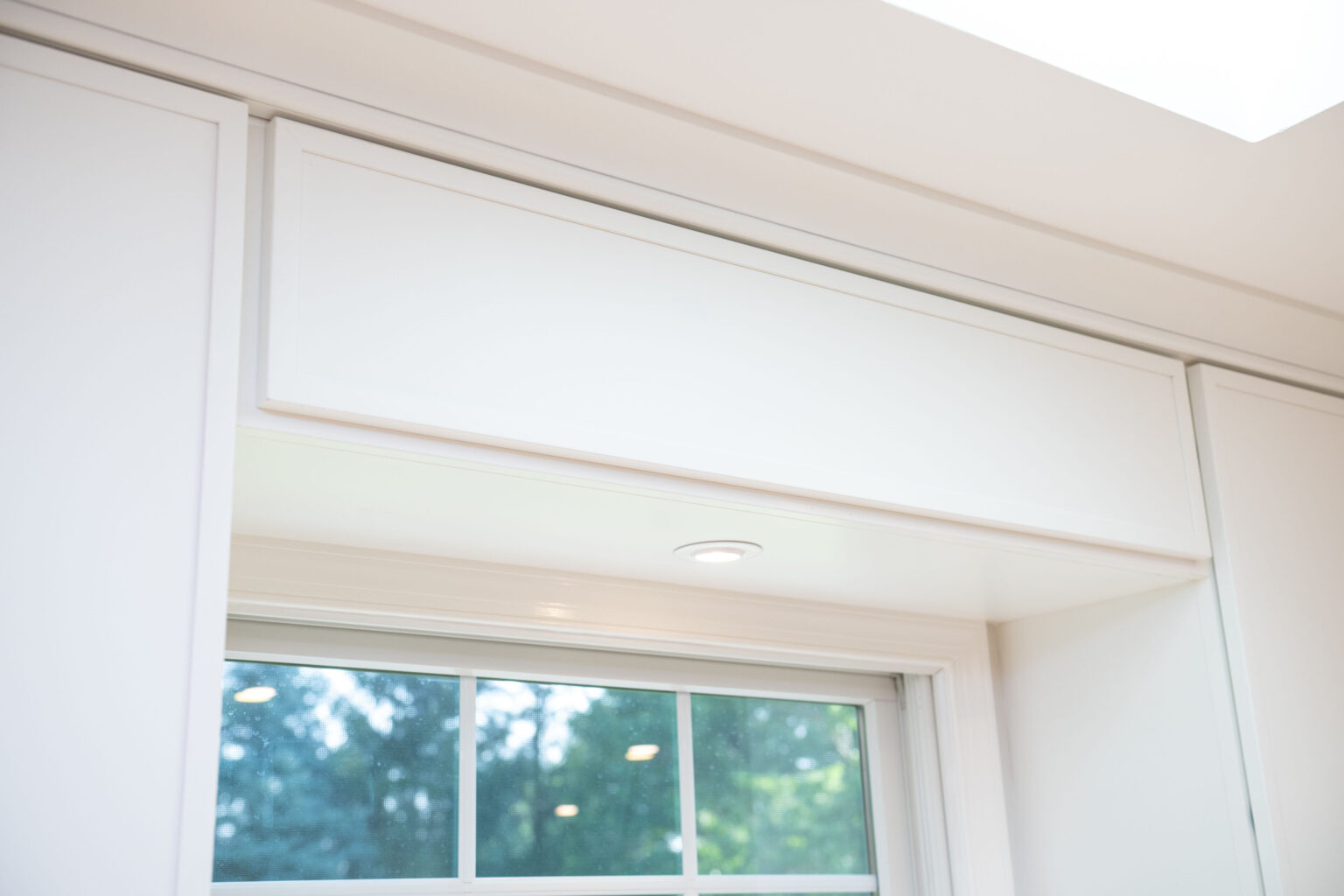 White kitchen cabinet with an overhead spotlight above a window, looking out onto green trees.