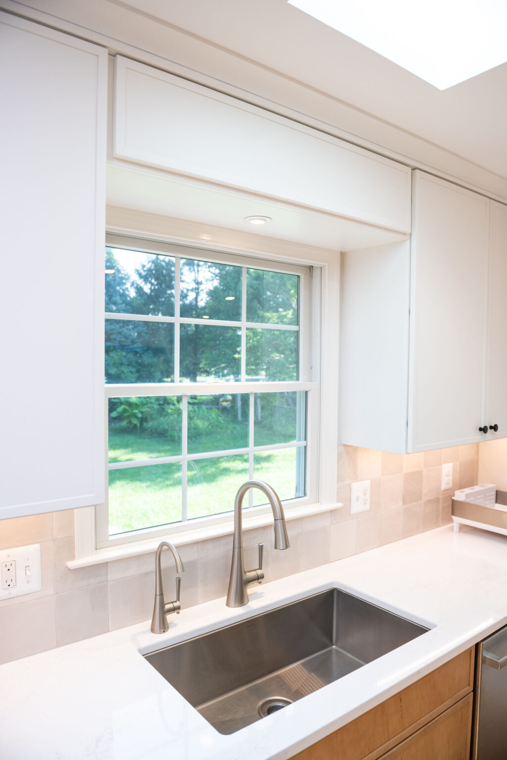 A modern kitchen sink with dual faucets set in a countertop below a window, overlooking a grassy yard. White cabinets and beige tile backsplash are visible.