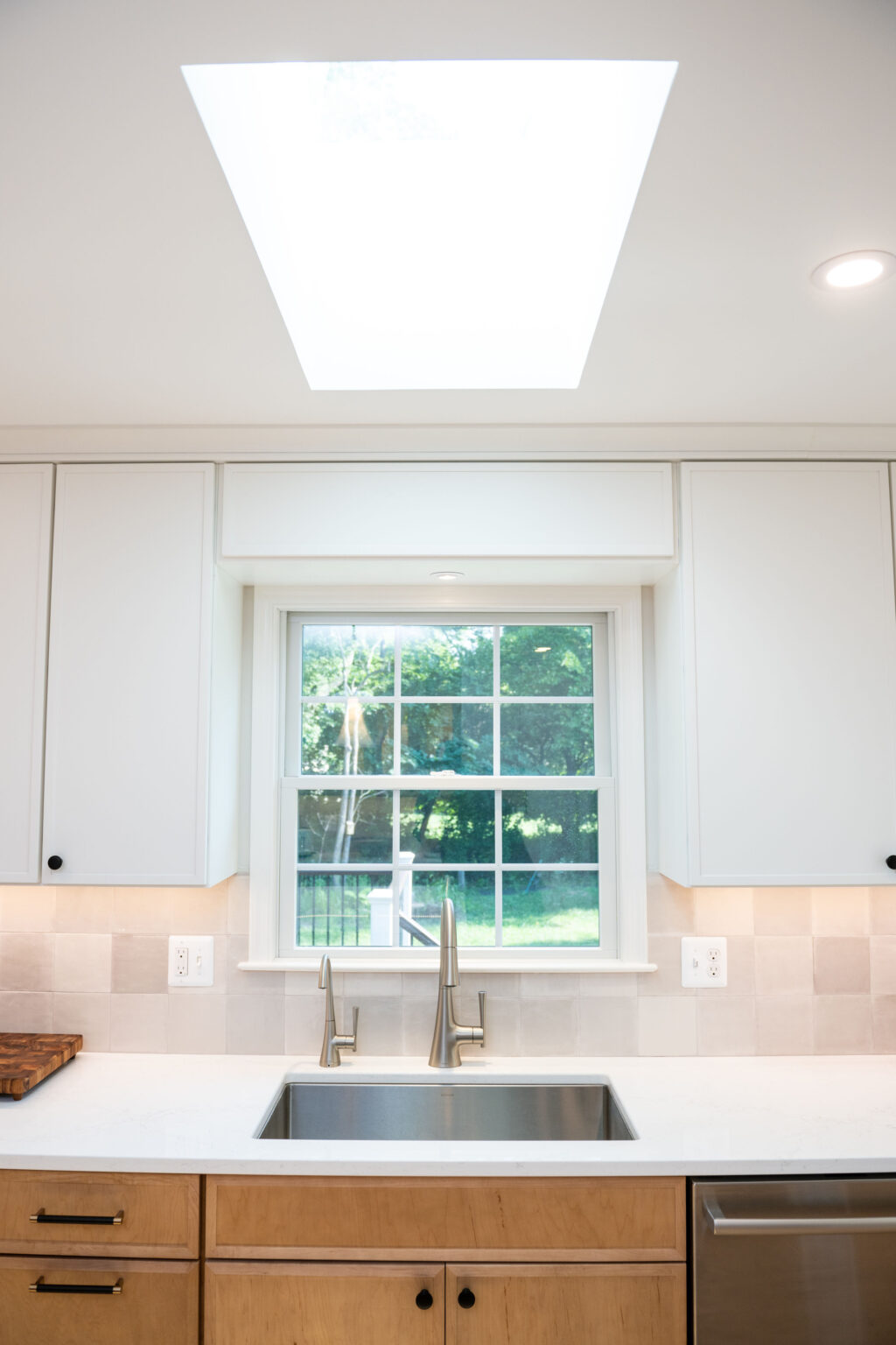 A modern kitchen features a stainless steel sink, light wood cabinets, and a window with a view of greenery, illuminated by a skylight.