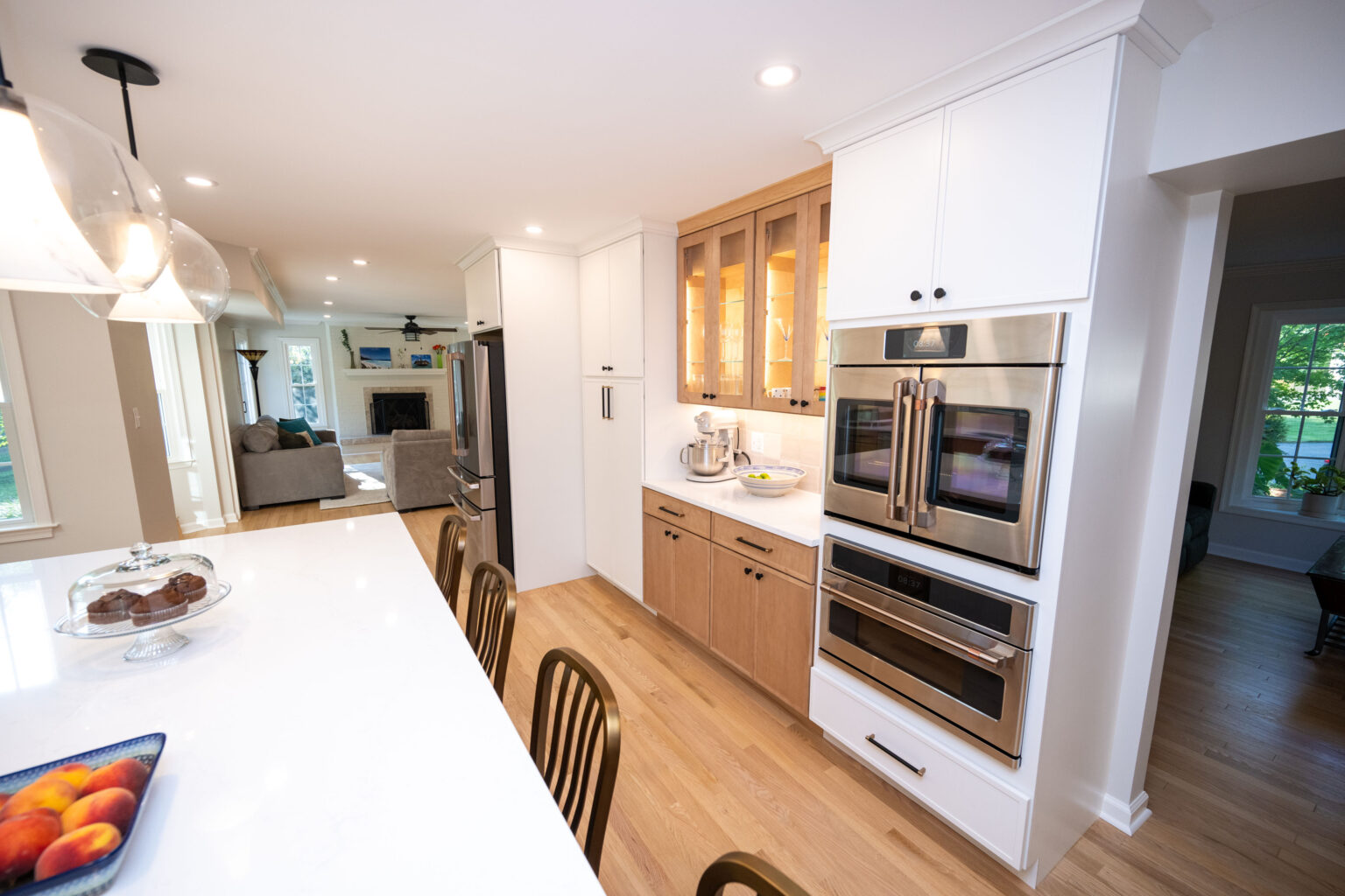 Modern kitchen with a white countertop, built-in double ovens, and a glass cabinet. Barstools line the island. A fruit bowl and cake stand are on the counter. Living area visible in the background.