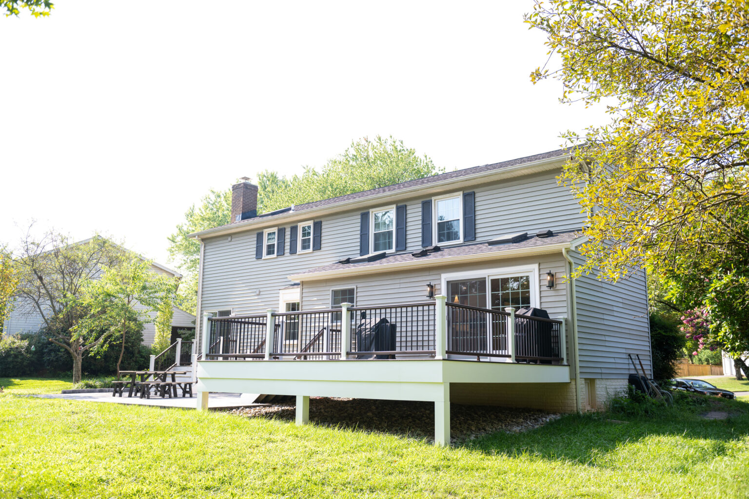A two-story house with gray siding features a raised deck in the backyard, surrounded by green grass and trees.