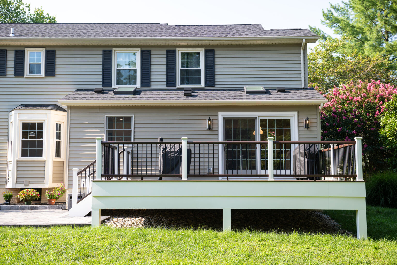 A two-story house with beige siding and black shutters features a wooden deck with railing in the backyard. The yard has green grass and a row of shrubs and flowers along the house.