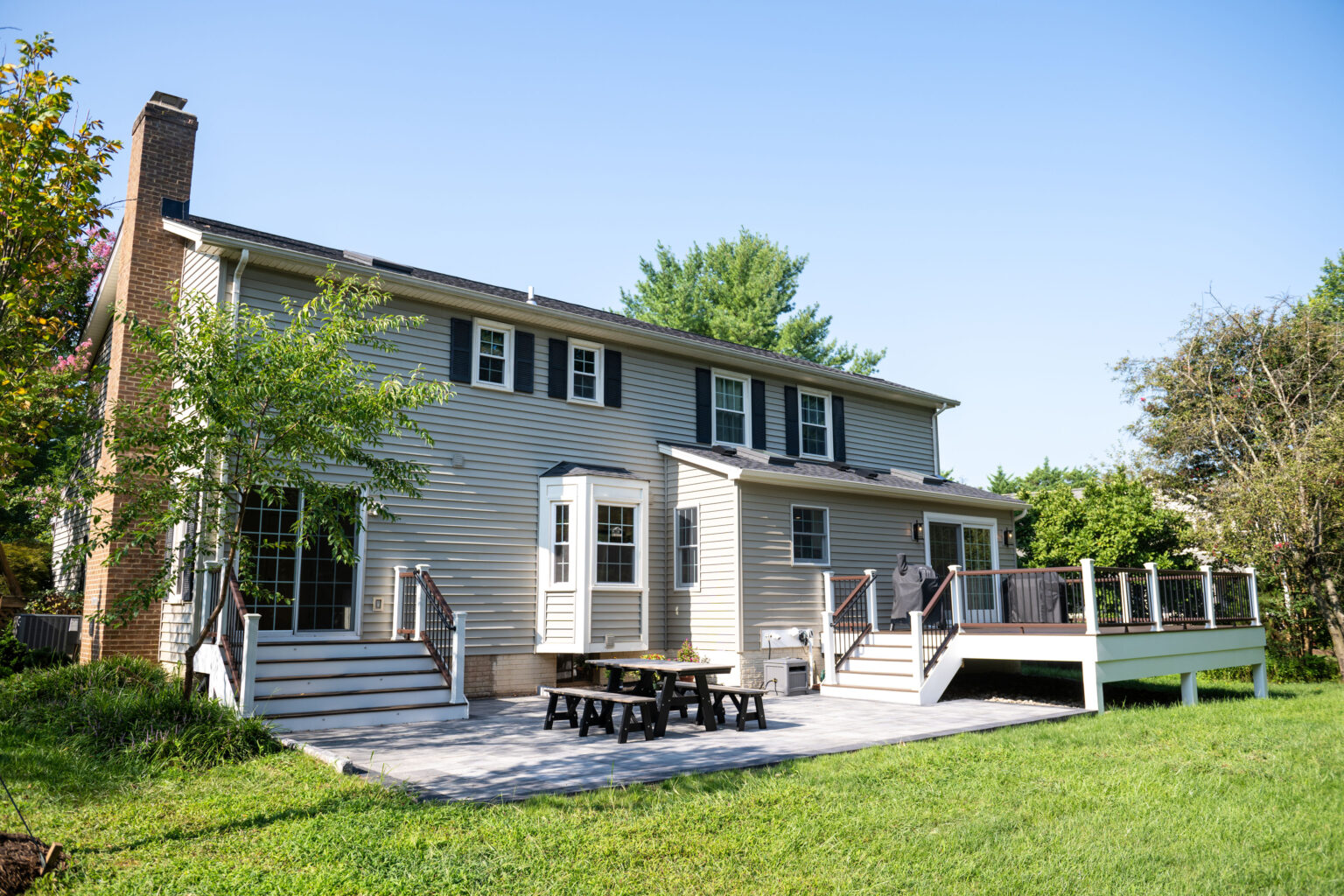 Two-story house with a patio, picnic table, and dual deck area surrounded by grass and trees under a clear blue sky.