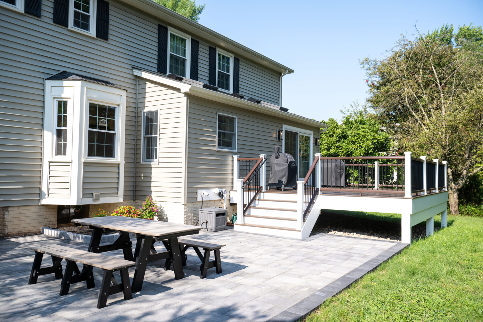 Two-story house with beige siding, a raised deck with railing, and a lower patio featuring a picnic table set. Trees and a grassy lawn are in the background.