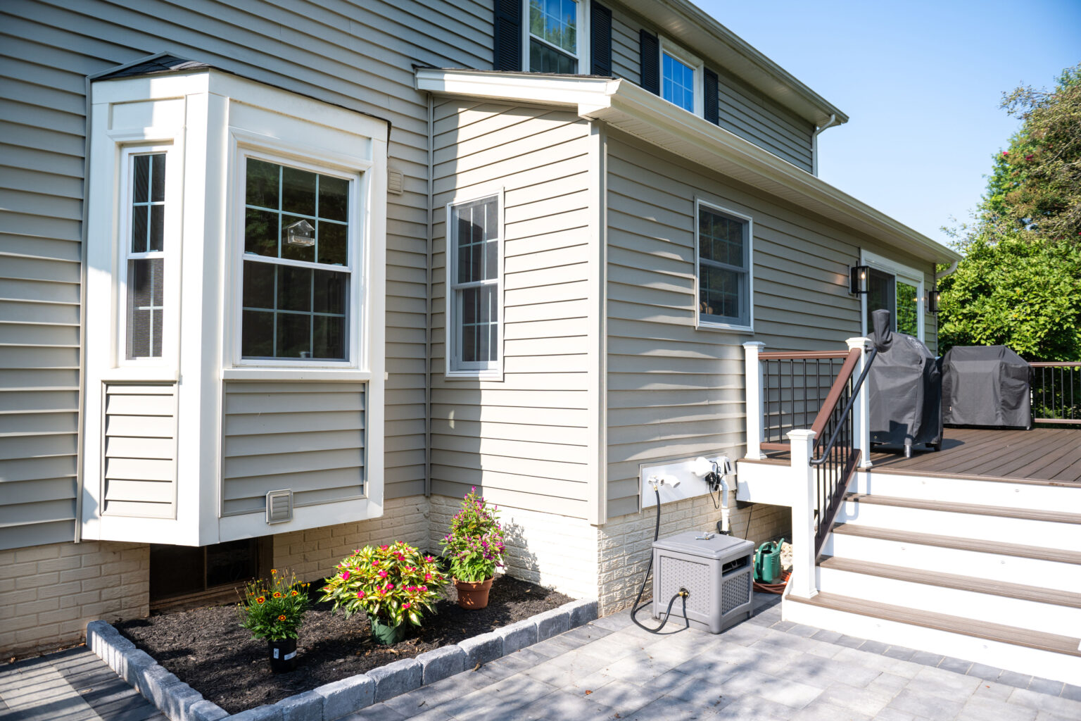 Backyard view of a house with beige siding, a bay window, stairs leading to a deck with covered grills, and a small garden bed with two potted plants.