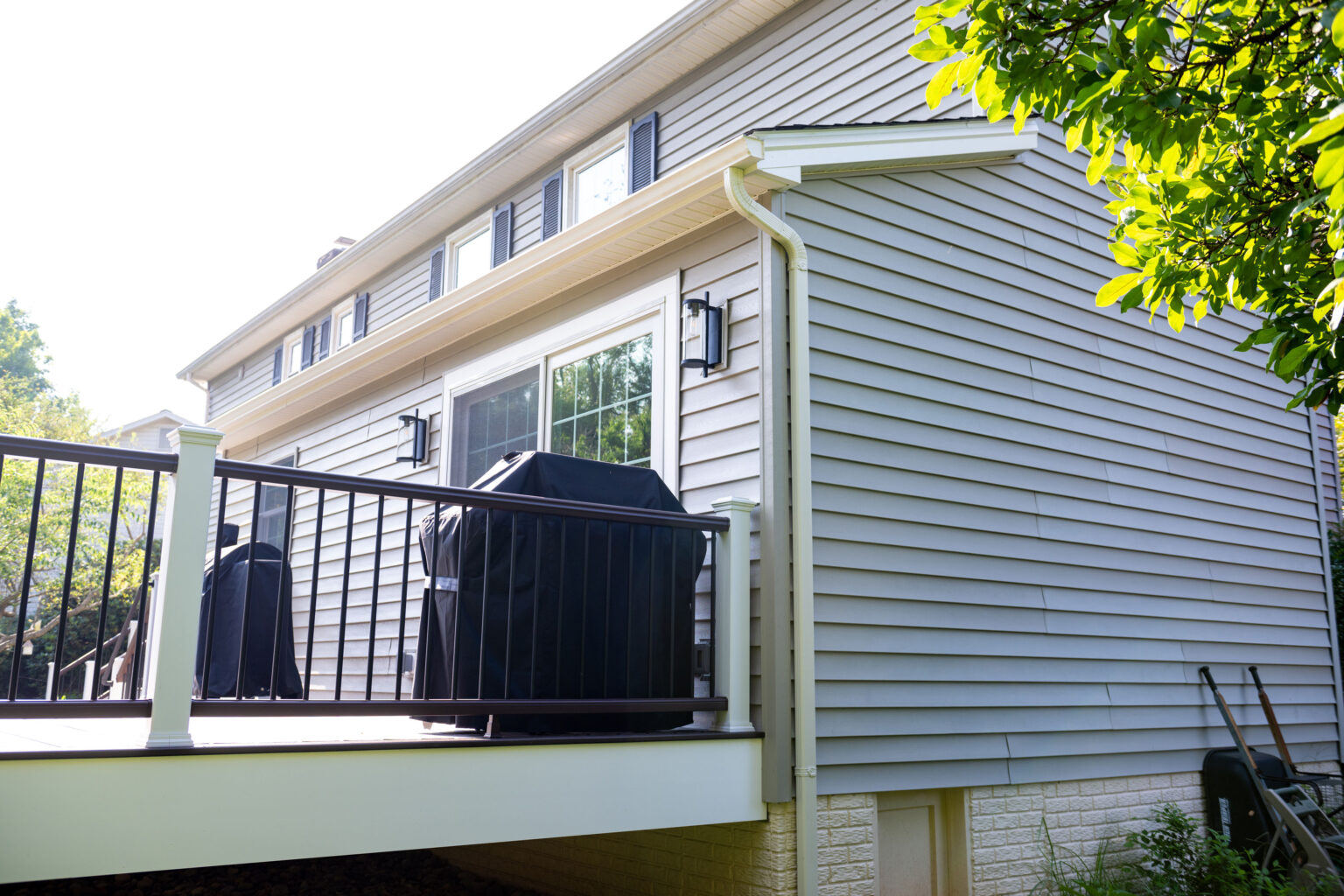A residential house with light gray siding and a deck featuring covered grills and black railing.