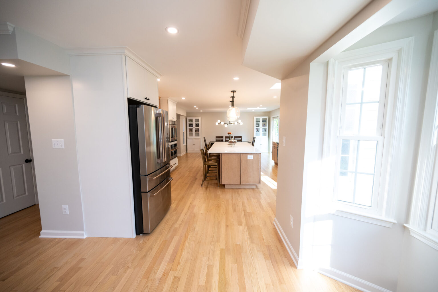 A spacious kitchen with light wooden flooring, a large central island, modern appliances, and pendant lights. Natural light streams in from a bay window.