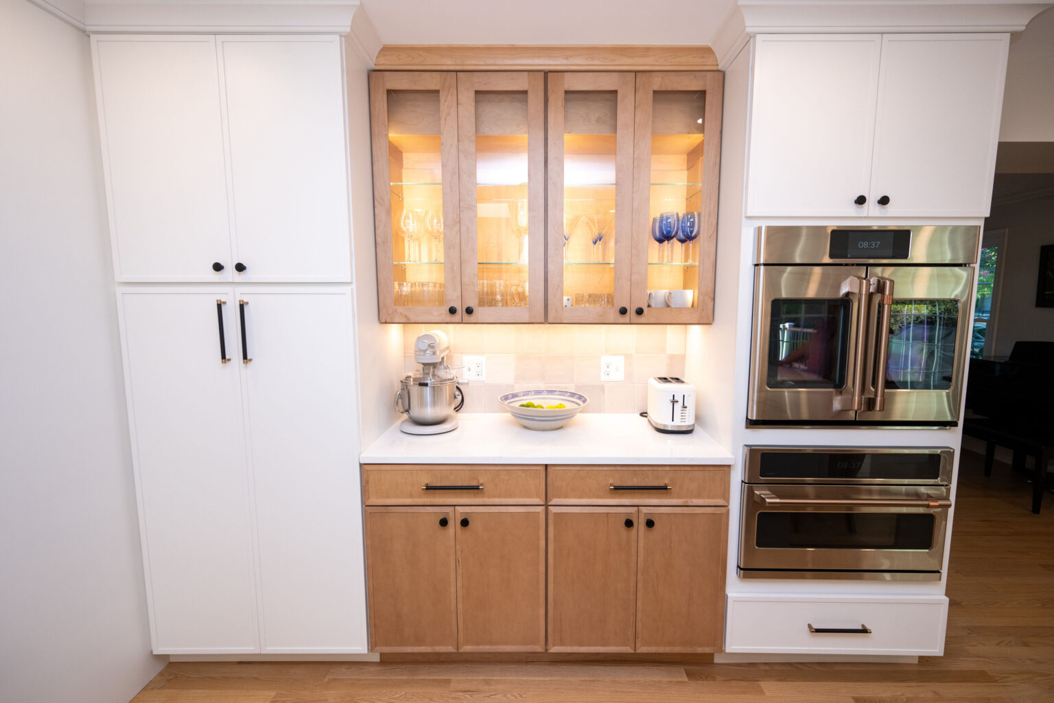 Kitchen with white and wood cabinets, a glass display, a mixer, toaster, bowl of lemons, and built-in double oven.