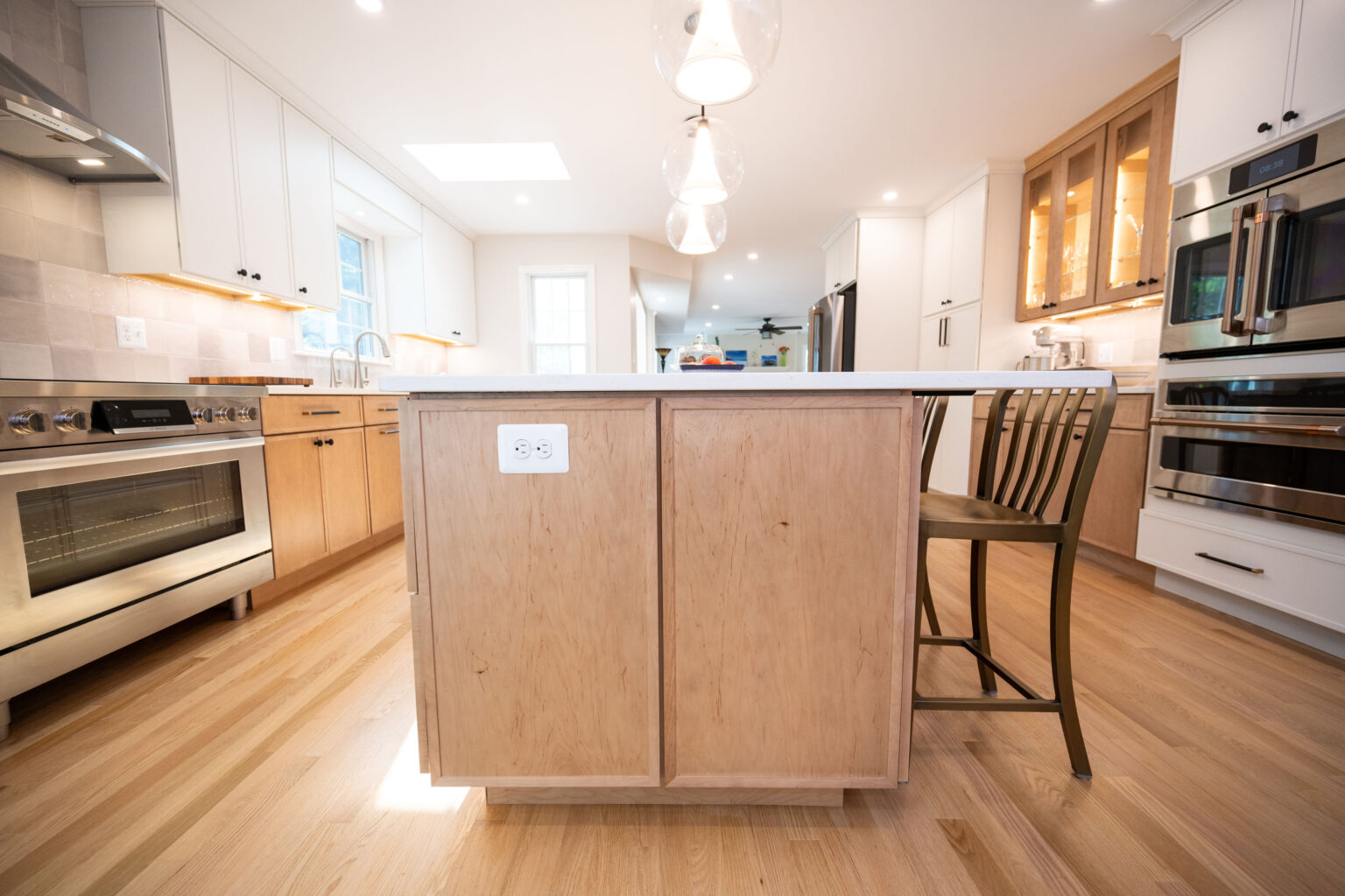 A modern kitchen with a large island in the center, featuring light wood cabinets, stainless steel appliances, and pendant lights. A chair is placed at the island.